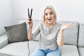 Poster - Young caucasian woman at home holding hair straightener celebrating achievement with happy smile and winner expression with raised hand