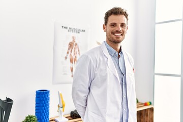 Young hispanic man wearing physiotherapist uniform standing at clinic