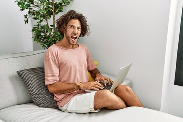 Wall Mural - Young hispanic man sitting on the sofa at home using laptop angry and mad screaming frustrated and furious, shouting with anger. rage and aggressive concept.