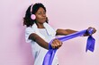 Young african american woman training arm resistance with elastic arm bands using headphones in shock face, looking skeptical and sarcastic, surprised with open mouth