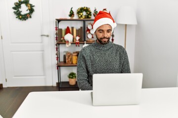 Canvas Print - Young hispanic man with beard wearing christmas hat using laptop with serious expression on face. simple and natural looking at the camera.