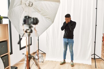 Wall Mural - Young hispanic man with beard posing as model at photography studio with sad expression covering face with hands while crying. depression concept.