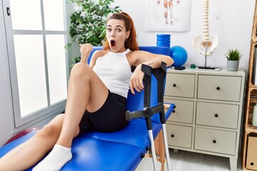 Poster - Young redhead woman lying on rehabilitation bed holding crutches surprised pointing with hand finger to the side, open mouth amazed expression.