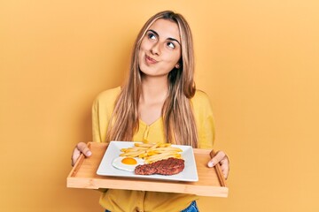 Poster - Beautiful hispanic woman holding tray with meat loaf and fried egg smiling looking to the side and staring away thinking.