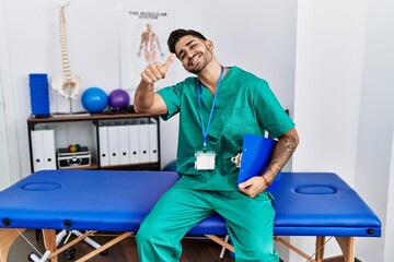 Sticker - Young physiotherapist man working at pain recovery clinic approving doing positive gesture with hand, thumbs up smiling and happy for success. winner gesture.