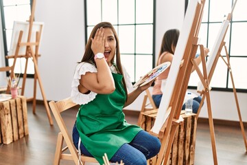 Sticker - Young hispanic artist women painting on canvas at art studio covering one eye with hand, confident smile on face and surprise emotion.