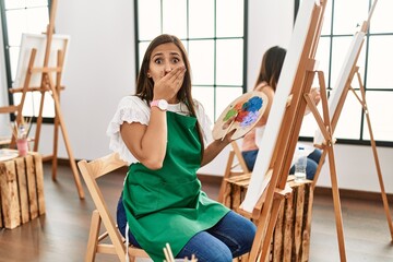 Poster - Young hispanic artist women painting on canvas at art studio shocked covering mouth with hands for mistake. secret concept.