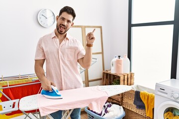 Poster - Young man with beard ironing clothes at home with a big smile on face, pointing with hand finger to the side looking at the camera.