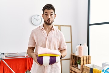 Poster - Young man with beard holding folded laundry after ironing making fish face with mouth and squinting eyes, crazy and comical.