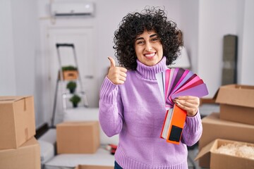 Canvas Print - Young brunette woman with curly hair choosing color of new house wall smiling happy and positive, thumb up doing excellent and approval sign