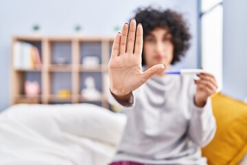 Sticker - Young brunette woman with curly hair holding pregnancy test result with open hand doing stop sign with serious and confident expression, defense gesture