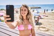 Young blonde girl make selfie by the smartphone sitting on the bench at the beach.