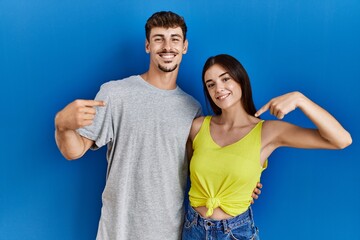 Poster - Young hispanic couple standing together over blue background looking confident with smile on face, pointing oneself with fingers proud and happy.