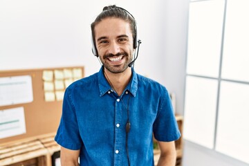 Young hispanic man with beard wearing call center agent headset at the office looking positive and happy standing and smiling with a confident smile showing teeth