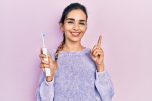 Young Hispanic Girl Holding Electric Toothbrush Smiling With An Idea Or Question Pointing Finger With Happy Face, Number One