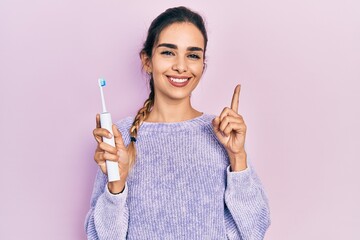 Wall Mural - Young hispanic girl holding electric toothbrush smiling with an idea or question pointing finger with happy face, number one