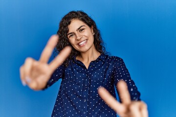 Poster - Young brunette woman with curly hair wearing casual clothes over blue background smiling looking to the camera showing fingers doing victory sign. number two.