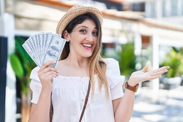 Sticker - Young brunette woman holding dollars banknotes celebrating achievement with happy smile and winner expression with raised hand