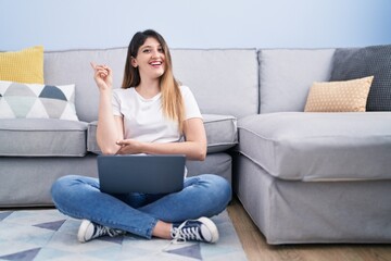 Sticker - Young brunette woman sitting on the floor at home using laptop smiling happy pointing with hand and finger to the side