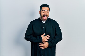 Canvas Print - Handsome hispanic man with beard wearing catholic priest robe smiling and laughing hard out loud because funny crazy joke with hands on body.