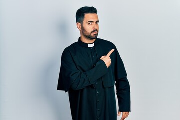 Sticker - Handsome hispanic man with beard wearing catholic priest robe pointing with hand finger to the side showing advertisement, serious and calm face