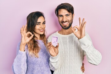 Beautiful couple of boyfriend and girlfriend holding engagement ring for proposal doing ok sign with fingers, smiling friendly gesturing excellent symbol