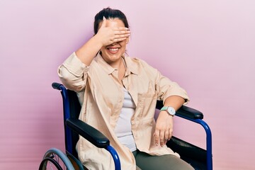 Poster - Young hispanic woman sitting on wheelchair smiling and laughing with hand on face covering eyes for surprise. blind concept.