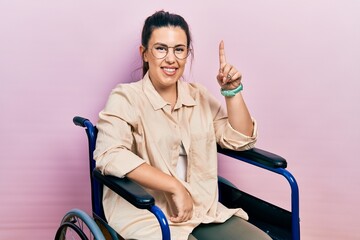 Sticker - Young hispanic woman sitting on wheelchair pointing finger up with successful idea. exited and happy. number one.