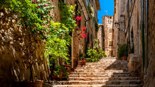 Romantic Narrow Alley With Rising Stairs Lined With Red Flowers And Potted Plants In Front Of Typical Mediterranean Stone Houses In The Center Of The Picturesque Mountain Village Fornalutx, Mallorca.