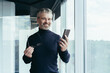 © Liubomir - Portrait of a successful gray-haired man, senior businessman boss in the office smiling and looking at the camera by the window, holding a phone and glasses