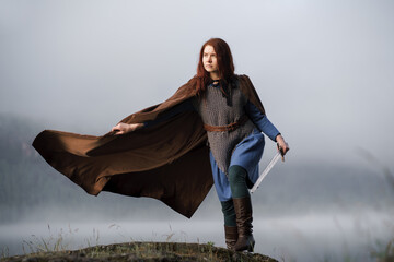 Wall Mural - Red-haired girl in armor with sword and raincoat on river bank