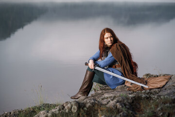 Wall Mural - Red-haired girl in armor with sword and raincoat on river bank