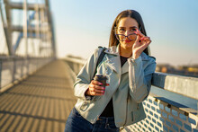Happy Beautiful Young Woman Enjoying In Sunset, She's Walking On City Bridge While Holding Phone And Cup Of Coffee And Posing For Photo Shoot.