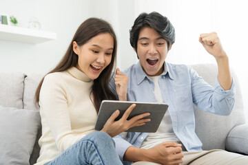 Happy excited, smiling asian young couple love using tablet pc, great deal or business success, received or getting cash back, tax refund, good news by mail while sitting on sofa, couch at home.