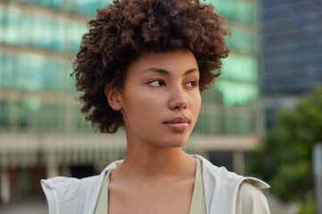 Wall Mural - outdoor shot of pensive curly haired young woman concentrated away strolls at street dressed in stre