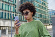 canvas print picture - Photo of stylish curly haired young woman wears sunglasses and green jumper checks received email message browses website in social media stands outdoors against modern buildings on urban street