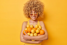 Indoor Shot Of Pensive Curly Haired Woman Wears Sunglasses Embraces Heap Of Citrus Fruits Eats Healhy Food Full Of Vitamins Poses Against Bright Yellow Background. Female Model With Oranges And Lemons