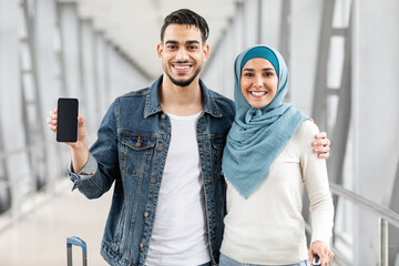 Wall Mural - Travel App. Happy Muslim Couple Standing At Airport And Showing Blank Smartphone