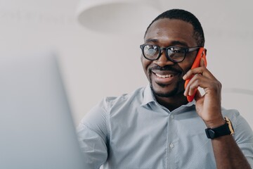 Happy african american businessperson in glasses hold phone makes business call in front of laptop