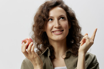 Wall Mural - Cheerful enjoyed curly beautiful woman in casual khaki green shirt hold apple point finger look up posing isolated on over white background. Natural Eco-friendly products concept. Copy space. Closeup