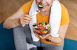 Sports and nutrition concept. Fit senior woman sitting on yoga mat, eating fresh vegetable salad after domestic workout