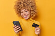 © wayhome.studio  - Positive curly haired European woman smiles happily uses modern smartphone and credit card purchases something makes shopping online dressed in casual jumper isolated over vivid yellow background