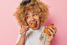 Beautiful Woman Feels Very Hungry Eats Chicken Nuggets With Ketchup Wears Dirty T Shirt And Protective Helmet Enjoys Harmful Fast Food Looks Away Isolated Over Pink Background. Unhealthy Eating