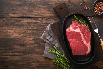 Wall Mural - Beef steak. Fresh raw beef rib eye steak with fork rosemary, salt and pepper on a grill pan on dark old wooden table background. Shop advertising idea. Top view. Mockup for design idea.