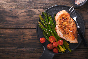 Wall Mural - Chicken breast grilled with vegetables. Grilled chicken steak, asparagus and cherry tomatoes in black plate on wooden dark table background. Healthy diet lunch. Barbecue steak fried. Top view.