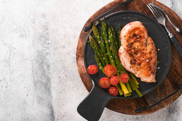 Wall Mural - Chicken breast grilled with vegetables. Grilled chicken steak, asparagus and cherry tomatoes in black plate on light gray concrete table background. Healthy diet lunch. Barbecue steak fried. Top view.