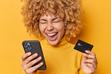 Overjoyed woman laughs out positively holds credit card and smartphone uses app for purchasing online dressed in casual jumper isolated over vivid yellow background. Modern technologies concept