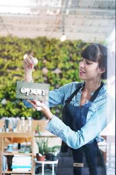 Smiling Caucasian Young barista woman or florist is wearing apron for setting open sign label on glass door in cafe or flowers shop. Start up for Coffee shop and cafe business Concept.