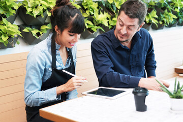 Smiling Caucasian Young barista couple love or partnership is wearing apron and consulting for marketing planning idea with tablet in the coffee shop. Start up for small cafe business owner concept.