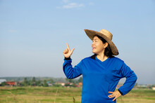 Portrait Of Asian Woman Farmer Wears Hat, Blue Shirt, Hand On Hip, Raise Finger To Point Up, Sky Background. Copy Space For Adding Text Or Advertisement. Concept : Agriculture Occupation. Happy Farmer